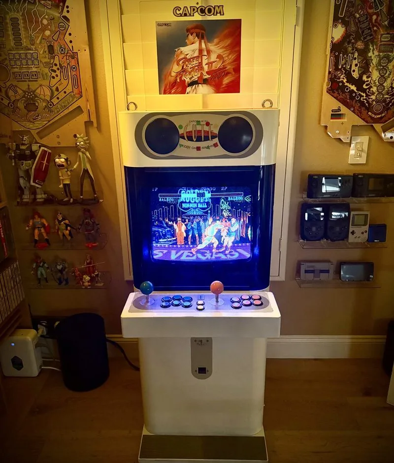 The pink Capcom Mini Cute candy cabinet head on at night, Street Fighter II lit on its screen and a Capcom poster above